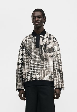 Mand iført en sort-hvid abstrakt mønstret langærmet polo og sorte bukser, stående foran en ensfarvet lys baggrund.