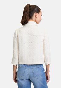 Witte tweedjas met driekwartmouwen, voorzien van een gestructureerd oppervlak en subtiele metalen deeltjes. Verkorte stijl met zijsplitten.