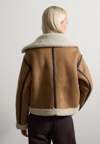 Chaqueta de ante marrón con collar de borrego, costuras y puños en negro. Presenta un corte cuadrado y un detalle de costura visible a lo largo de los bordes.