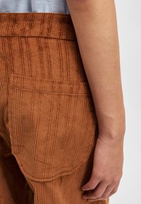 Calças de cordura de cor ferrugem com riscas verticais, apresentando um bolso traseiro e uma cintura elástica, destacando uma superfície suave e texturizada.