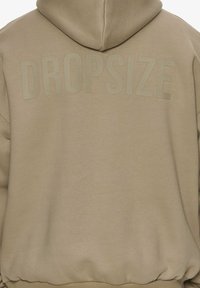 Beige hoodie med et stort, afdæmpet "DROPSIZE" print på ryggen. Blødt stof, afslappet pasform, og ribbede kanter og ærmer.