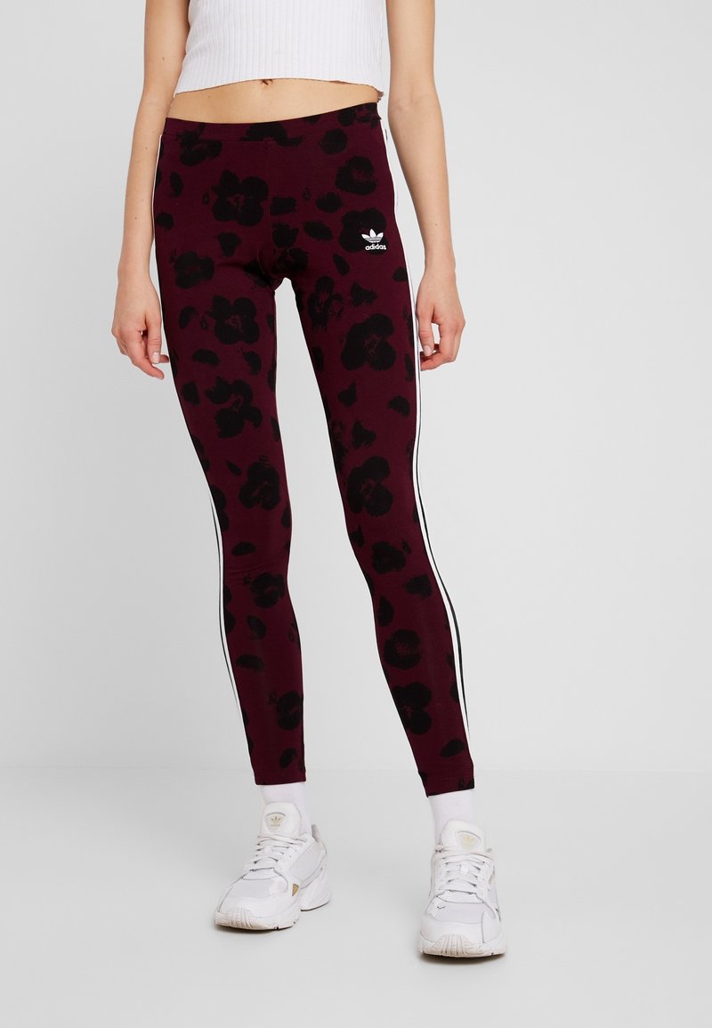Bordowe legginsy z czarnym kwiatowym wzorem, z białymi paskami po bokach i logo Adidas na pasku. Elastyczny materiał i dopasowany krój.
