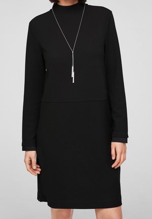Person wearing a long-sleeve black ribbed dress with a silver pendant necklace against a plain light gray background.