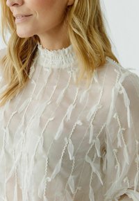 White sheer top featuring a high smocked collar, textured with delicate ruffles and vertical strands. Light fabric creates a flowing appearance.