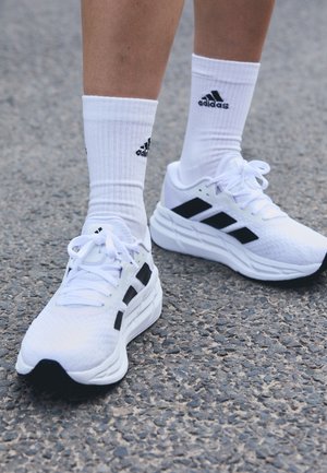 Jambes portant des chaussures de course Adidas blanches à rayures noires et des chaussettes hautes Adidas blanches, debout sur un bitume texturé.