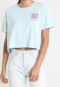 Světle modré zkrácené tričko vyrobené z bavlny, s čtvercovým purpurovým logem a textem "HOME BOY" na levé straně hrudi.