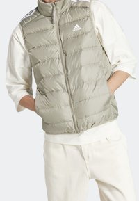 Světle zelená pufovaná vesta s vysokým límcem, zipovým zapínáním a vodorovným prošíváním; má bílé logo Adidas a kontrastní pruhy na ramenech.