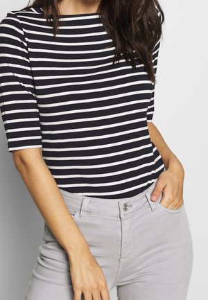 Femme portant une chemise rayée noir et blanc avec des manches jusqu'au coude et un pantalon gris clair, une main dans la poche, sur un fond uni.