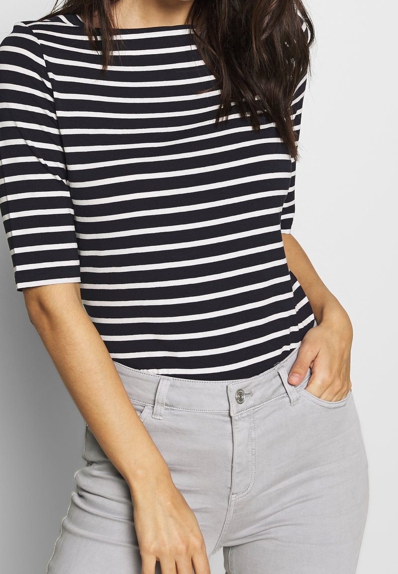 Femme portant une chemise rayée noir et blanc avec des manches jusqu'au coude et un pantalon gris clair, une main dans la poche, sur un fond uni.