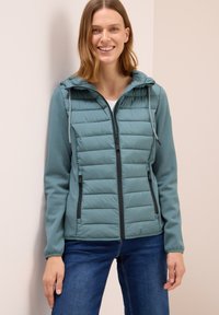 Kvinde iført en teal-farvet quiltet zip-up jakke med hætte og blå jeans, som smiler og læner sig op ad en beige væg.