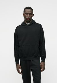 Must värvikapühi, mis on valmistatud pehmest kangast, ees olevate taskute, ribidega mansettide ja alumise osa korral, koos sobivate tumedate pükstega.