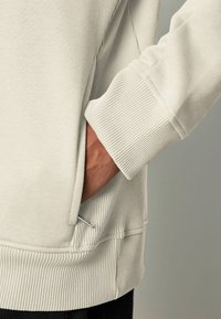 Sudadera gris claro con puño acanalado, bolsillo lateral y tejido texturizado. El bolsillo tiene un pequeño detalle plateado.