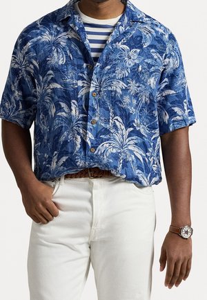 Mężczyzna ubrany w niebieską koszulę na guziki z krótkim rękawem w tropikalny wzór, nałożoną na granatowo-rampersowy t-shirt, białe spodnie, brązowy pleciony pasek oraz zegarek na nadgarstek z brązowym paskiem.