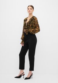 Zwarte op maat gemaakte broek gecombineerd met een doorschijnende blouse met bloemenpatroon en ruffles. Model draagt zwarte slingback hakken.