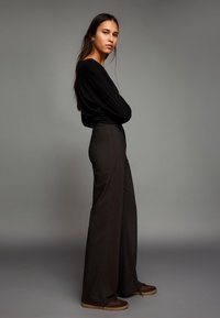 Bluza neagră cu mâneci lungi, asortată cu pantaloni largi cu dungi închise la culoare, cu talie înaltă. Încălțăminte maro, stând pe un fundal neutru.