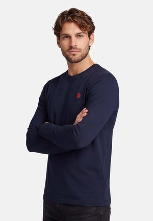 Mann mit hellbraunen Haaren und Bart, trägt ein dunkelblaues Langarmhemd mit kleinem rotem Logo auf der Brust, Arme verschränkt.