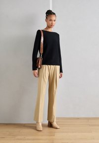 Schwarzes Langarmoberteil, beigetönige, plissierte Hose und hellfarbige Absatzschuhe. Braune, strukturierte Handtasche mit Schulterriemen.