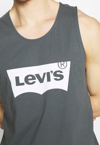 Grå bomullslinne med en framträdande vit Levi's-logotyp i en stiliserad, böjd design tvärs över bröstet. Slät textur, avslappnad passform.