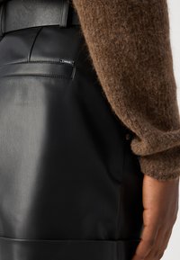 Gros plan d'une personne portant un pantalon en cuir noir avec une manche de pull marron en peluche et une main reposant sur le côté.