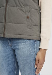 Grau isolierte Weste mit Reißverschluss und einer Druckknopftasche, kombiniert mit hellblauen Jeans. Weiche Textur und dezente Stickdetails sind sichtbar.