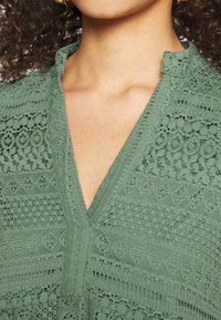 Green textured blouse featuring intricate lace patterns, a v-neckline, and a subtle collar. The fabric has a soft, woven appearance.