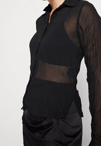 Vrouw in een zwarte sheer blouse met lange mouwen, gedragen over een zwarte bralette, gecombineerd met een zwarte satijnen gerimpelde rok.