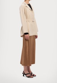 Beige Blazer mit Gürtel und Reverskragen, kombiniert mit einen braunen Satinrock und schwarzen Sandalen mit runder Ferse.