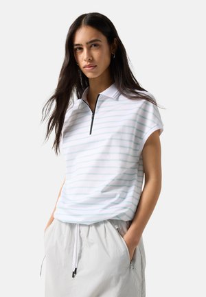 Młoda kobieta o długich ciemnych włosach, ubrana w białe polo z krótkim rękawem z jasnymi paskami i białe spodnie ze sznurkiem, ręce w kieszeniach.