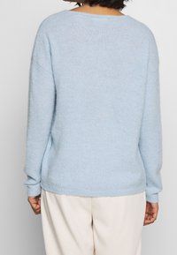 Jasnoniebieski sweter z dzianiny, miękka tekstura, luźny krój, szerokie rękawy, zaokrąglony dekolt, boczne rozcięcia przy dolnym brzegu, noszony z kremowymi spodniami.