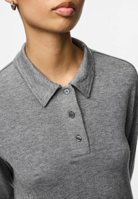 Detailní záběr na osobu, která má na sobě šedý pletený polo svetr se třemi knoflíky a malým kruhovým náušnicem na bílém pozadí.