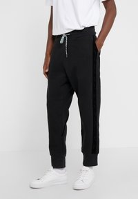 Persona vestida con pantalones jogger negros con cintura ajustable y rayas laterales, combinados con zapatillas blancas y calcetines blancos, de pie frente a un fondo blanco.