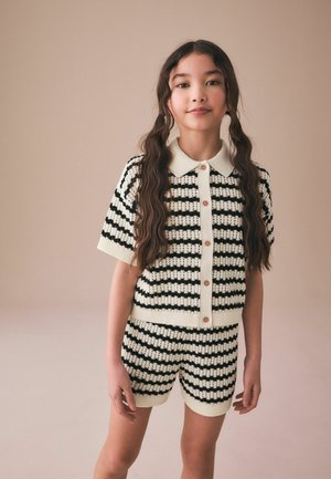 Noor tüdruk, kellel on pikad lainelised juuksed, kannab musta-valge triibulist soonikkoes nööpidega pusa ja sobivaid lühikesi pükse, seistes beež tausta ees.