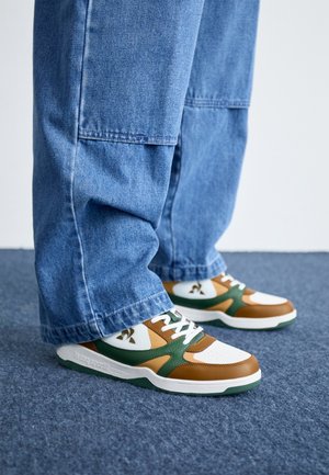 Persoon die blauwe wijde jeans en bruine, witte en groene sneakers draagt, staand op een blauw tapijt.