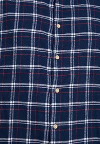 Camicia di flanella a quadri navy con motivi a scacchi rossi e bianchi, dotata di cinque bottoni beige lungo la parte anteriore. Texture morbida.