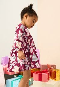 Lila klänning med långärmade och en utsvängd kjol, med mönster av djur och julrelaterade föremål. Omgiven av färgglada presentlådor.