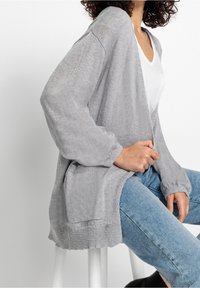 Světle šedý pletený cardigan s dlouhými rukávy, s otevřeným předním dílem a dvěma bočními kapsami, překrytý bílým topem a modrými džínami. Jemná textura.