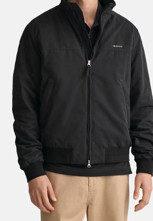 Mann trägt eine schwarze Jacke mit Reißverschluss, gerippten Bündchen und einem kleinen "GANT"-Logo auf der Brust, kombiniert mit beigen Hosen.