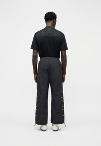 Άνδρας που στέκεται με την πλάτη γυρισμένη, φορώντας μαύρο κοντομάνικο πουκάμισο, μαύρο παντελόνι με φαρδιά μπατζάκια και ρίγες στο πλάι, και λευκά αθλητικά παπούτσια σε λευκό φόντο.