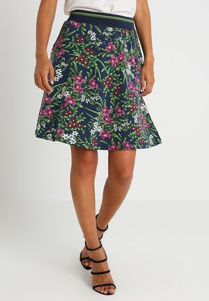 Femme portant une jupe patineuse bleu marine à imprimé floral rose, blanc et vert, associée à des sandales noires à talons hauts et fines brides.