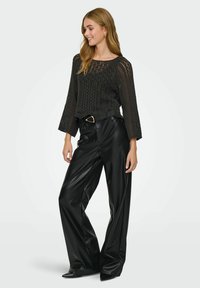 Femme aux longs cheveux portant un pull en crochet noir, un pantalon en cuir noir à jambes larges, une ceinture avec une boucle en or et des chaussures noires à bouts pointus, souriant.