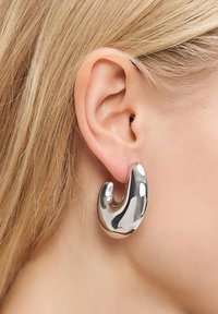 Närbild på en blond persons öra som bär en slät, glänsande, silverfärgad abstrakt hoopsä kring.