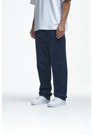 Homme portant un jean bleu foncé ample, des baskets blanches et un t-shirt blanc oversized, debout sur un sol blanc.