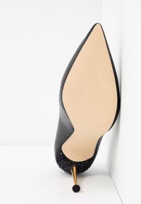 Escarpin noir à bout pointu avec semelle beige, adhérence texturée, "GUESS" embossé, et contrefort scintillant sur une surface blanche.