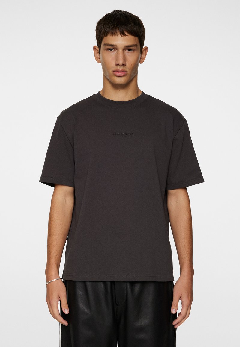 Mörkgrå bomull t-shirt med avslappnad passform, kort ärm och en subtil tryckt logotyp på bröstet. Slät textur och rundad halsdesign.