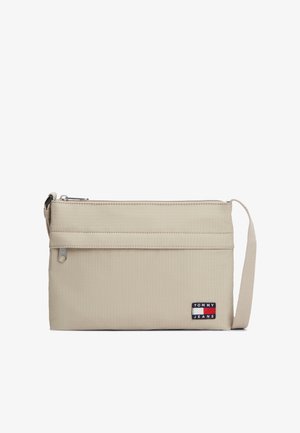 Sac bandoulière beige en tissu texturé, doté d'une fermeture éclair, d'une sangle ajustable, et d'un patch logo Tommy Jeans visible.