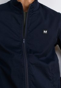 Granatowa kurtka na zamek wykonana z lekkiego, gładkiego materiału, z wysokim kołnierzem i małą, prostokątną naszywką z logo na lewej piersi.