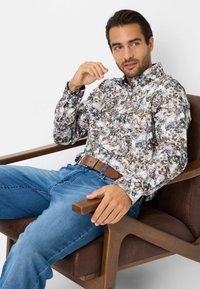 Camicia a maniche lunghe con motivo floreale nei toni del bianco, blu e marrone, abbinata a jeans di denim blu e una cintura di pelle marrone, indossata mentre si è seduti.