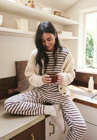 Gestreiftes braunes und cremefarbenes Loungewear-Set kombiniert mit einem beigen Cardigan auf einer Küchenarbeitsplatte. Halten eine braune Keramiktasse, mit einem Holz-Hintergrund.