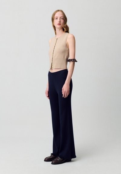 Top sin mangas cortado en beige con botones dorados, combinado con pantalones anchos de canalé en azul marino oscuro y mocasines marrones con acentos de volantes.