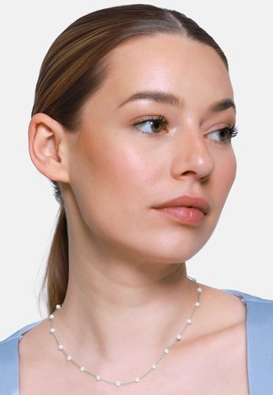 Giovane donna con pelle liscia e capelli castani raccolti, che indossa una delicata collana d'argento con piccole perle distribuite uniformemente e una maglietta azzurro chiaro.